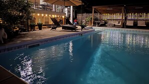 Una piscina con sillas de descanso y sombrillas, rodeada por una terraza de madera y un edificio con guirnaldas de luces.
