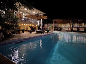 Une piscine entourée d’une terrasse en bois et d’un bâtiment illuminé de guirlandes lumineuses, avec des chaises longues et des parasols à proximité.
