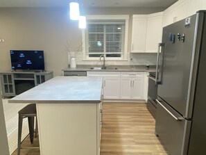 A modern kitchen with white cabinets, a stainless steel refrigerator, a built-in oven, and a flat-screen TV. - Vedder River Retreat near UFV, Cultus Lake, Shops, Restaurants, Garrison. (Chilliwack)