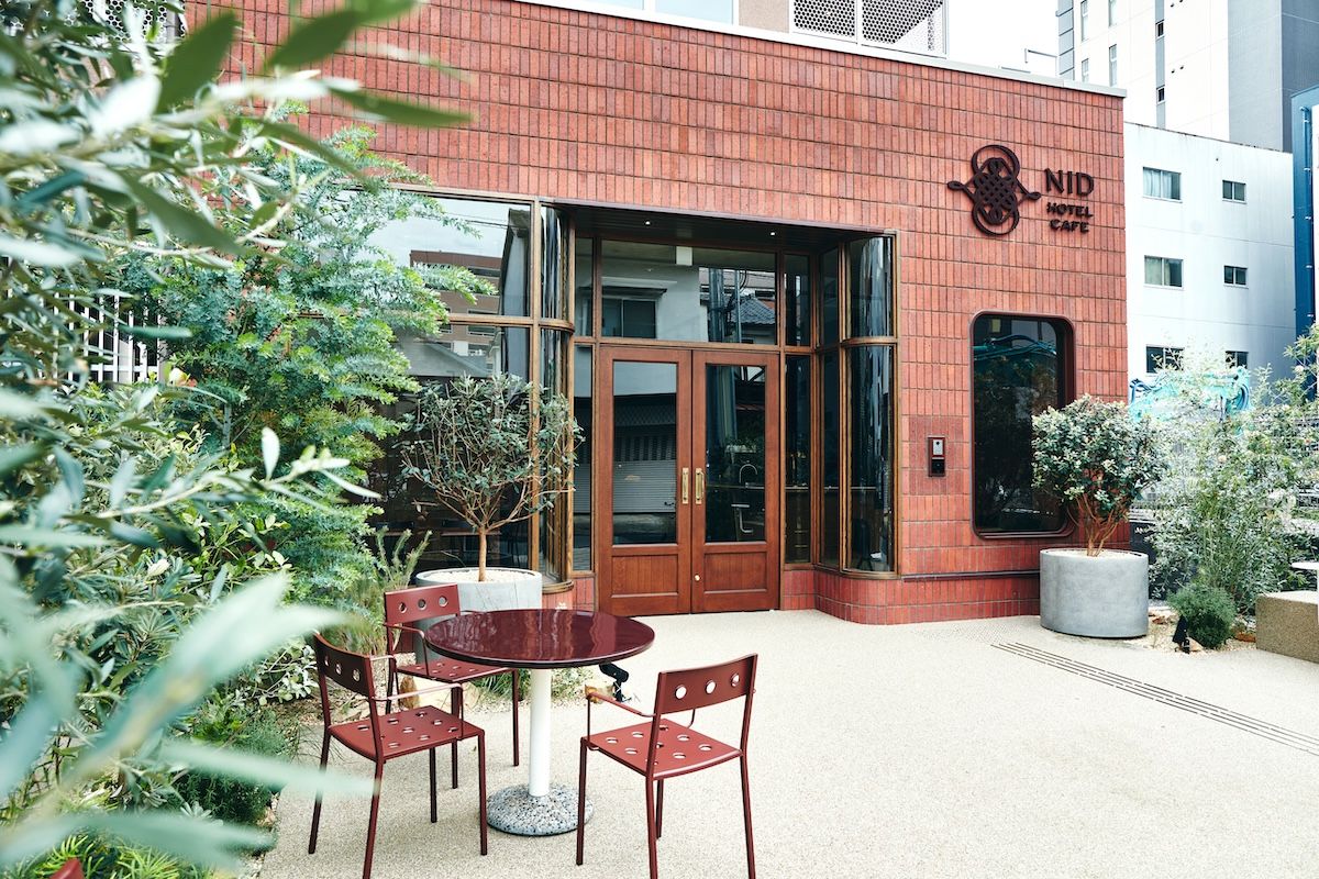 Entrada de hotel com fachada de tijolos vermelhos, porta de vidro e um pequeno espaço ao ar livre com uma mesa redonda e cadeiras.