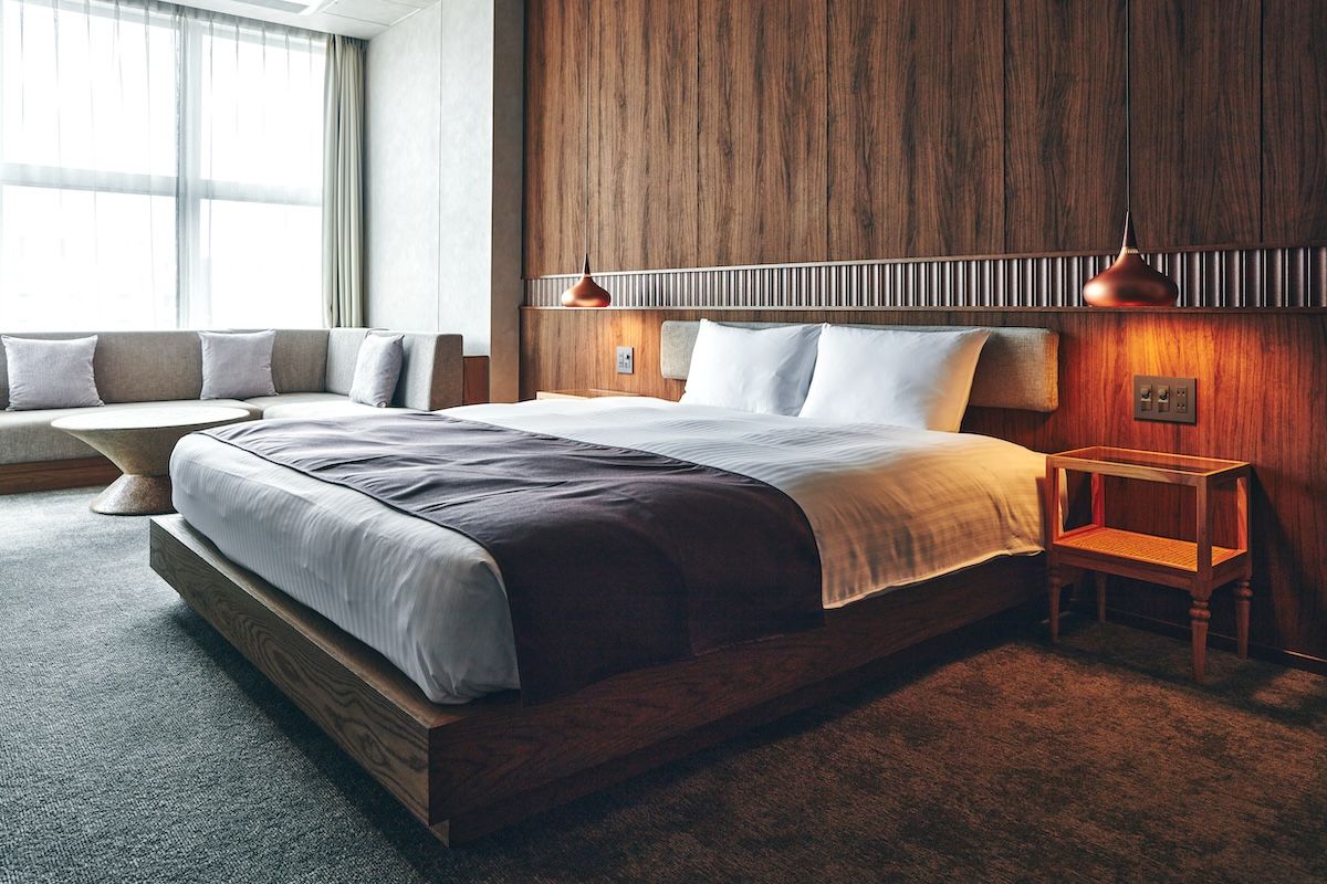 Quarto moderno com cabeceira de madeira, cama grande, mesa de cabeceira e um espaço de estar com um banco.