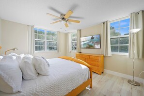 Ein Schlafzimmer mit einem Bett, einem Deckenventilator, einer Kommode, einem Fernseher und großen Fenstern.