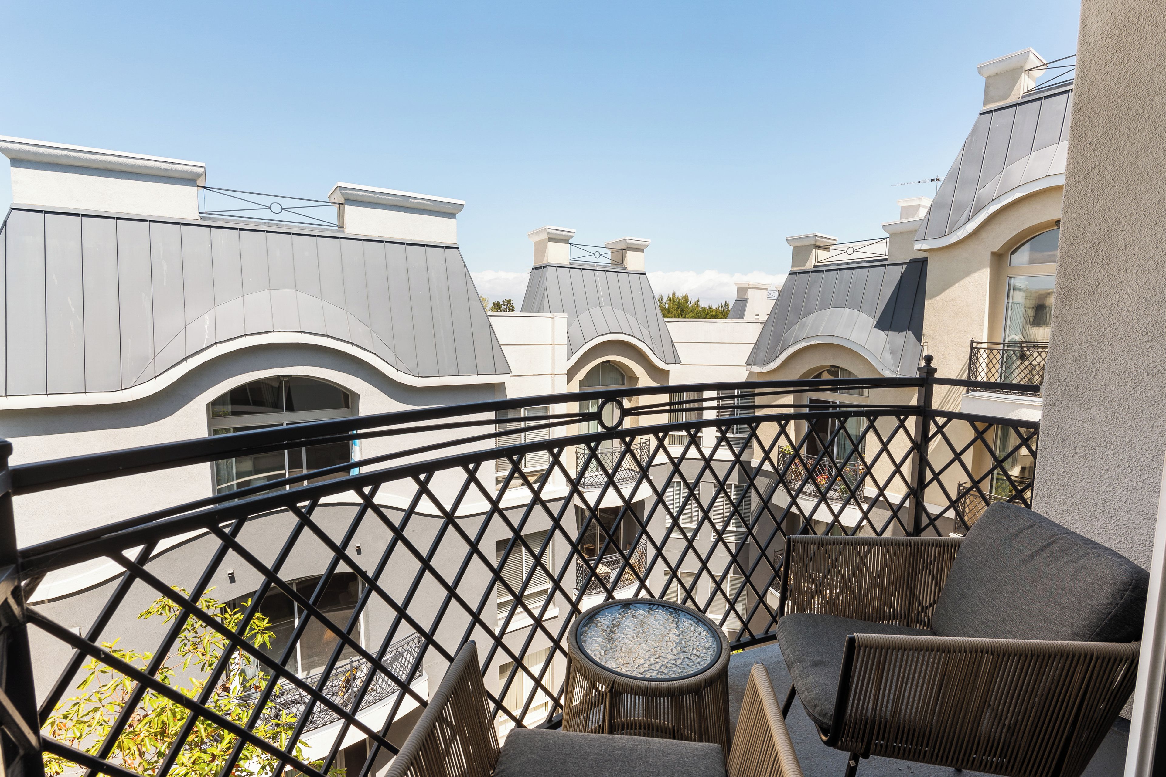 A balcony with wicker furniture and a small table, overlooking a residential area with unique architectural rooftops.