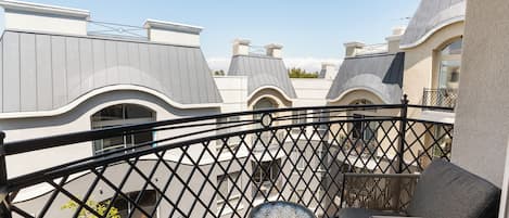 A balcony with wicker furniture and a small table, overlooking a residential area with unique architectural rooftops.