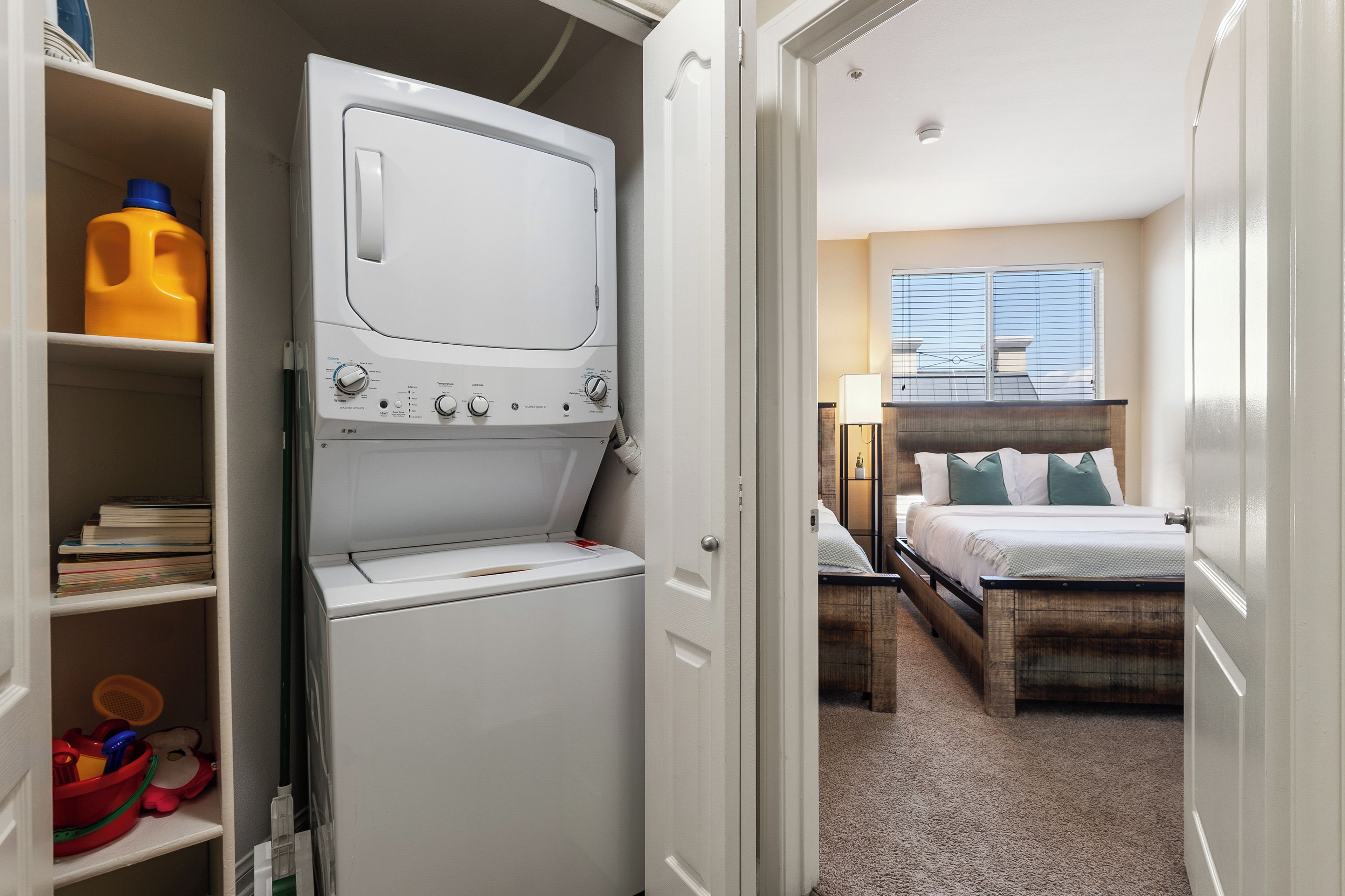 Ein kleines Hotelzimmer mit integrierter Waschmaschine und Trockner, einem Bett mit Holz Kopfteil, einem Nachttisch mit Lampe sowie einem Fenster mit Jalousien.