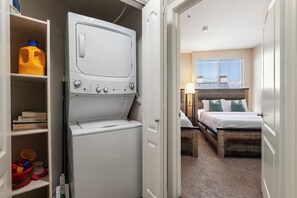 A compact hotel room with a built-in washer and dryer, a bed with a wooden headboard, a bedside table with a lamp, and a window with blinds. - Serene Zen' Quiet Garden View Resort Pool, Gym, Jacuzzi (Marina del Rey)