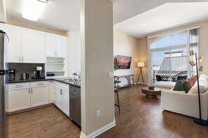 A modern kitchen with white cabinets, a coffee maker, and a sink. There is a dining area with a table and chairs, and a large window offering a view of the outdoors. - Serene Zen' Quiet Garden View Resort Pool, Gym, Jacuzzi (Marina del Rey)