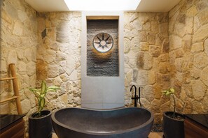 A stone-walled bathroom with a freestanding bathtub, a wall-mounted towel rack, and two potted plants. - Villa Santi Bayu by Alfred in Bali (Pecatu)