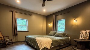 Quarto com cama, ventoinha no teto, duas janelas com cortinas, piso de madeira e uma pequena mesa com moldura de quadro.