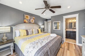 A bedroom with a bed, a ceiling fan, a painting, a nightstand, and a bedside table. - The Regency | Oceanfront | Private Pool (Virginia Beach)
