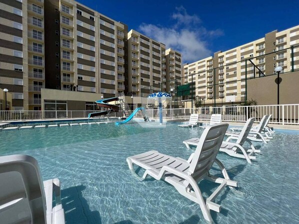 A swimming pool with lounge chairs, a water slide, and a building with multiple balconies in the background.
