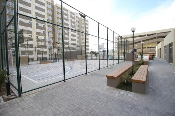 A fenced basketball court with a bench and a lamp post, surrounded by a paved walkway and a high-rise building in the background.
