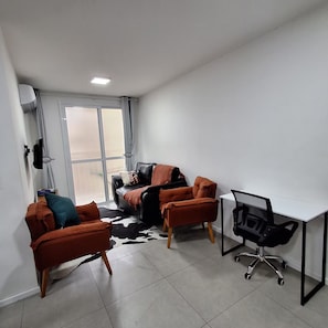 Un espace de bureau moderne avec un canapé en cuir noir, deux fauteuils orange, un bureau blanc avec une chaise noire et une grande fenêtre avec des rideaux.
