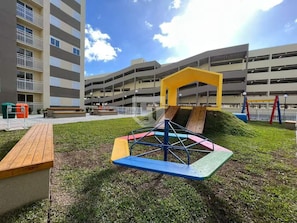 A playground with a slide and climbing structure, surrounded by a grassy area and a multi-story parking garage in the background.