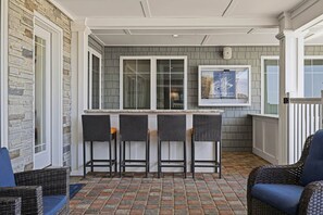 A covered outdoor area with a stone wall, a bar counter with stools, and wicker furniture. - The Regency Royale | Oceanfront | Private Pool (Virginia Beach)