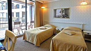 A hotel room with two beds, a desk, a chair, a balcony with a view of buildings, and a framed picture on the wall. - Regina Maria (Balchik)