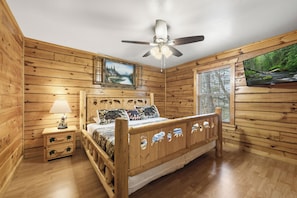 A wooden cabin room with a bed, a nightstand, a ceiling fan, a painting, and a window with a view of a forest. - Zen Bear’s Den our Beautiful cozy cabin 2 miles to Dollywood in Pigeon Forge TN. (Pigeon Forge)