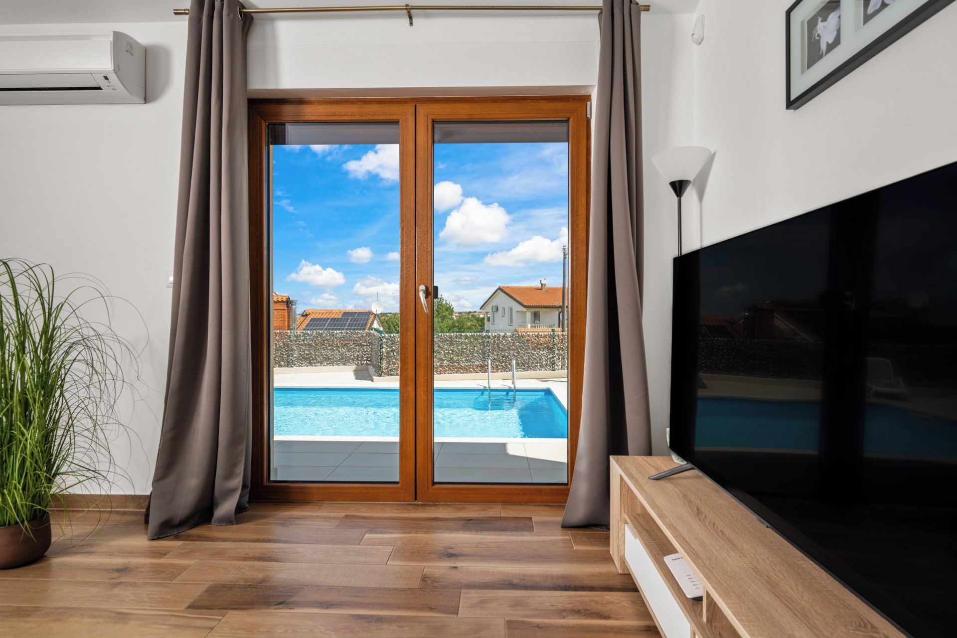A living room with a large TV, wooden flooring, a potted plant, and a view of a swimming pool through glass doors.