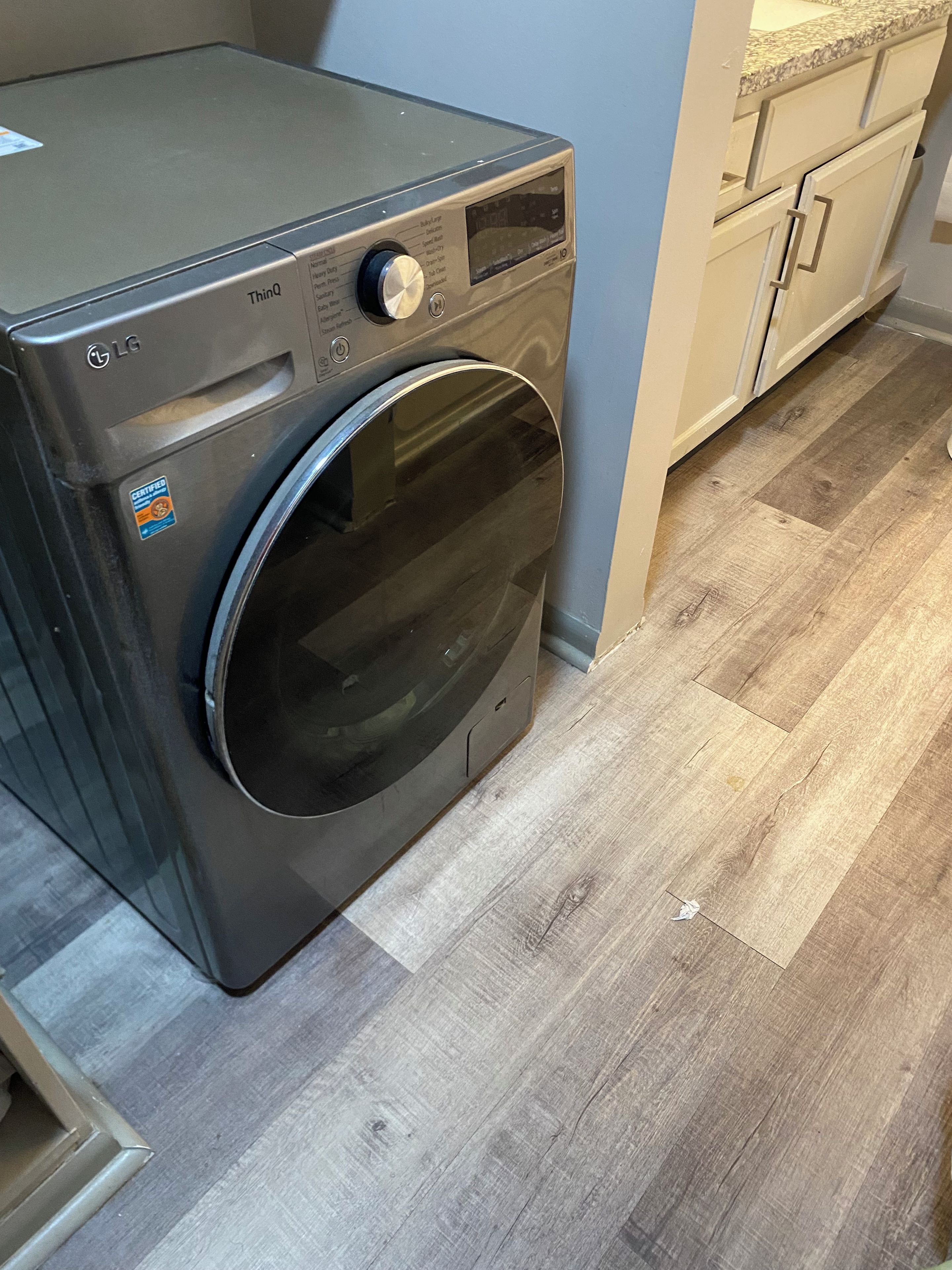A front-loading LG washer in a kitchen with wooden flooring and white cabinets.