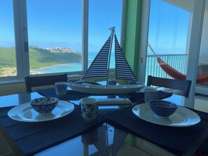 A dining area with a table set for two, a view of the ocean, and a hammock on the balcony. - Slow Down & Relax Ocean, Marina, and Mountain Views! (Fajardo)