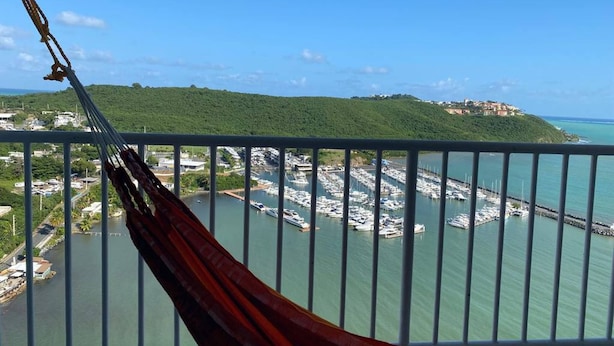 Uma rede em uma varanda com vista para um porto com barcos e uma paisagem montanhosa ao fundo.