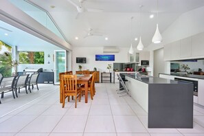 A modern kitchen with a central island, dining area with chairs, and a large window overlooking greenery. - Seaside Haven | 4 Bedroom Holiday Home with Private Pool (Palm Cove)