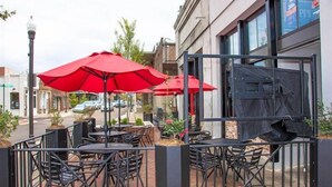 An outdoor seating area with tables and chairs, red umbrellas, and a metal fence. - "Rebel Depot" - By OVR (Oxford)
