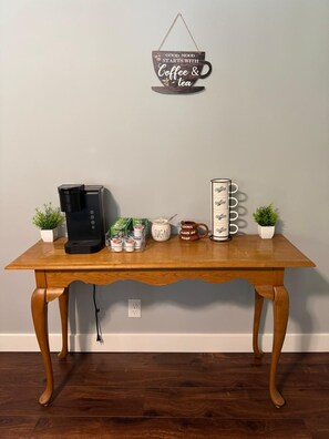 Ein Holztisch mit einer Kaffeemaschine, Zucker, Milchkartons, einer Tasse und einem Schild mit der Aufschrift 'Gute Laune beginnt mit Kaffee & Tee'.