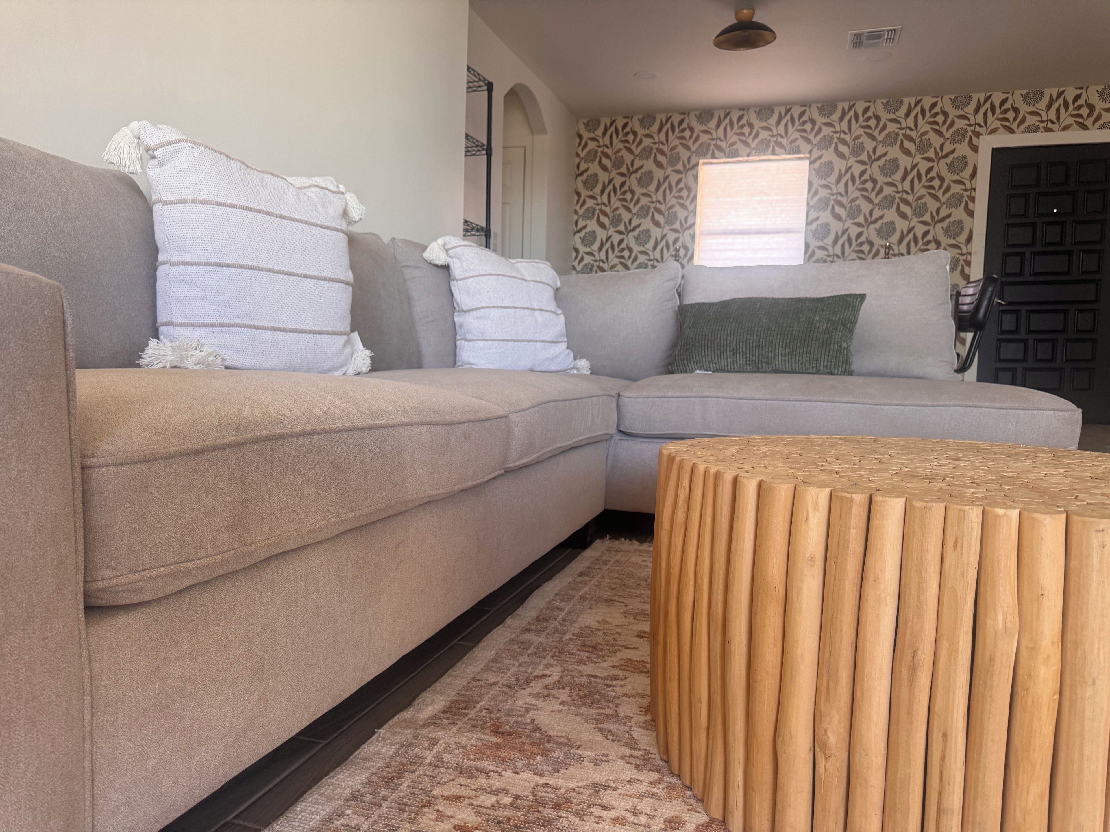 Un salon moderne avec un canapé d’angle beige, une table basse en bois et un papier peint à motifs.