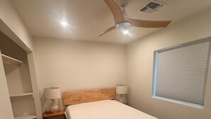 Ein Schlafzimmer mit einem Bett, einem Nachttisch, einem Deckenventilator, einem Fenster mit Jalousien und einem an der Wand befestigten Regal.