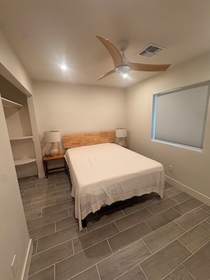 Un dormitorio con cama, mesita de noche, ventilador de techo, ventana con persianas y estante de pared.