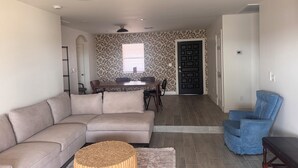 Ein Wohnzimmer mit einer Couch, einem Couchtisch, einem blauen Sessel, einem Essbereich mit Tisch und Stühlen sowie einer Wand mit Muster.
