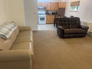 Sala de estar con sofá y sillón de cuero, área de cocina con gabinetes y electrodomésticos de madera, y una ventana con persianas.