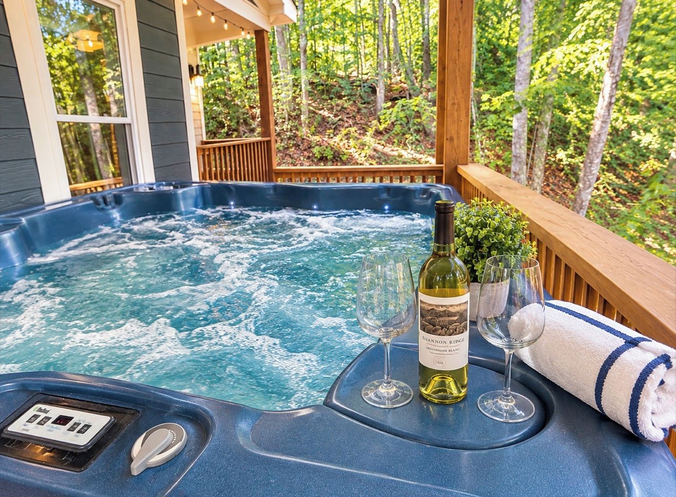 Een hottub met bedieningspaneel, twee wijnglazen, een fles wijn en een handdoek op een houten terras met uitzicht op een bos.