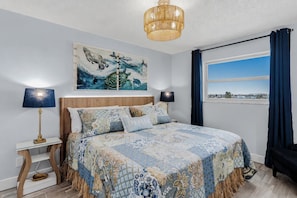 A bedroom with a bed, two bedside lamps, a ceiling light, a window with curtains, and a wall painting. - 🐠Sea Breeze 505: Gulf-front |Madeira Beach| Pool| Hot Tub| John's Pass! (Madeira Beach)