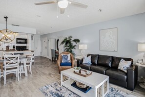 A modern living room with a black leather sofa, white coffee table, and a dining area with white chairs and a kitchen in the background. - 🐠Sea Breeze 505: Gulf-front |Madeira Beach| Pool| Hot Tub| John's Pass! (Madeira Beach)