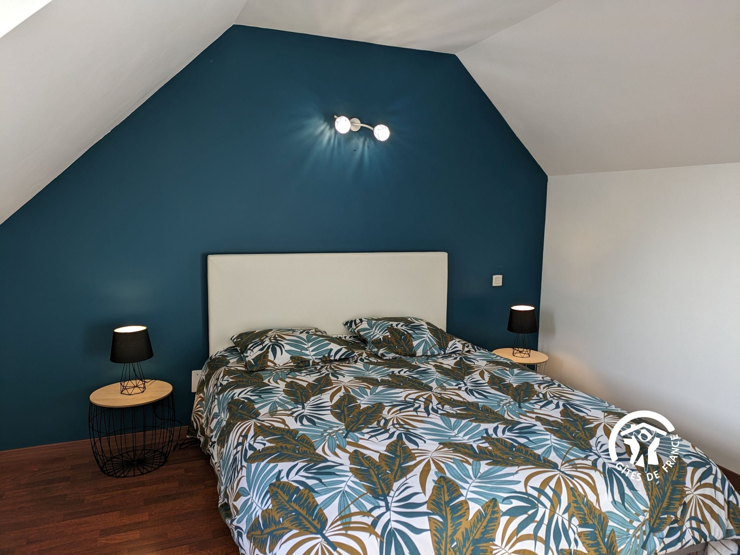 Un dormitorio con techo inclinado, una cama con colcha estampada de hojas, dos lámparas de noche y un aplique de pared.