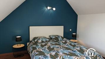 Un dormitorio con techo inclinado, una cama con colcha estampada de hojas, dos lámparas de noche y un aplique de pared.