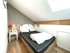 Habitació petita amb un llit individual, sostre inclinat, una taula de nit amb una làmpada i una finestra.