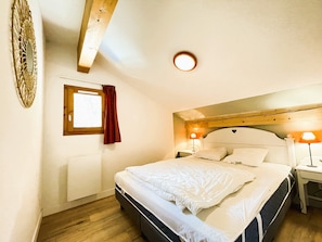 A bedroom with a bed, two bedside tables, a window with curtains, a wall-mounted light fixture, and a wooden ceiling beam. - Apartment with wifi, mountain view - 7 guests (Valfréjus)