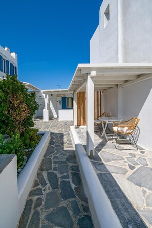 A white building with a stone pathway leading to a covered outdoor seating area with a table and chairs. - Gelos House (Mykonos)
