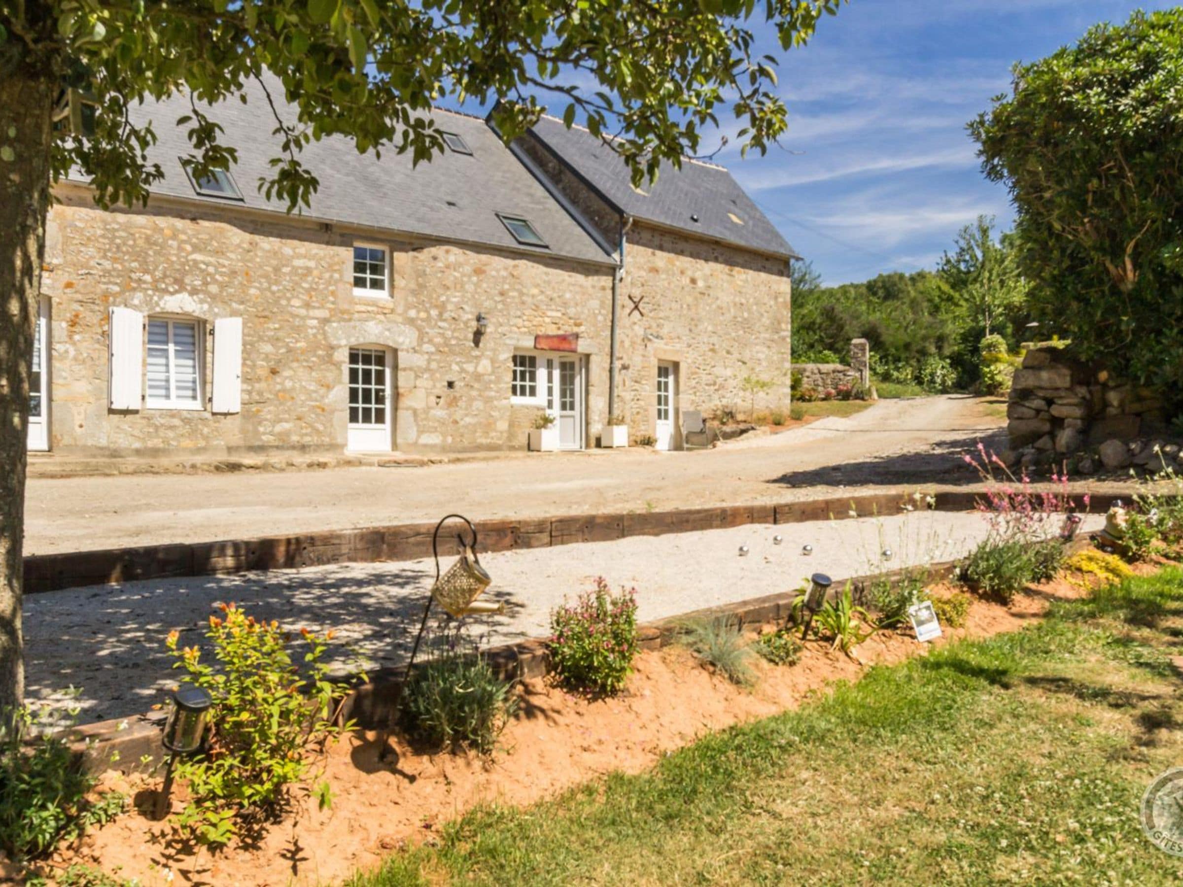 A stone building with a paved driveway and a garden with various plants and a bird feeder.
