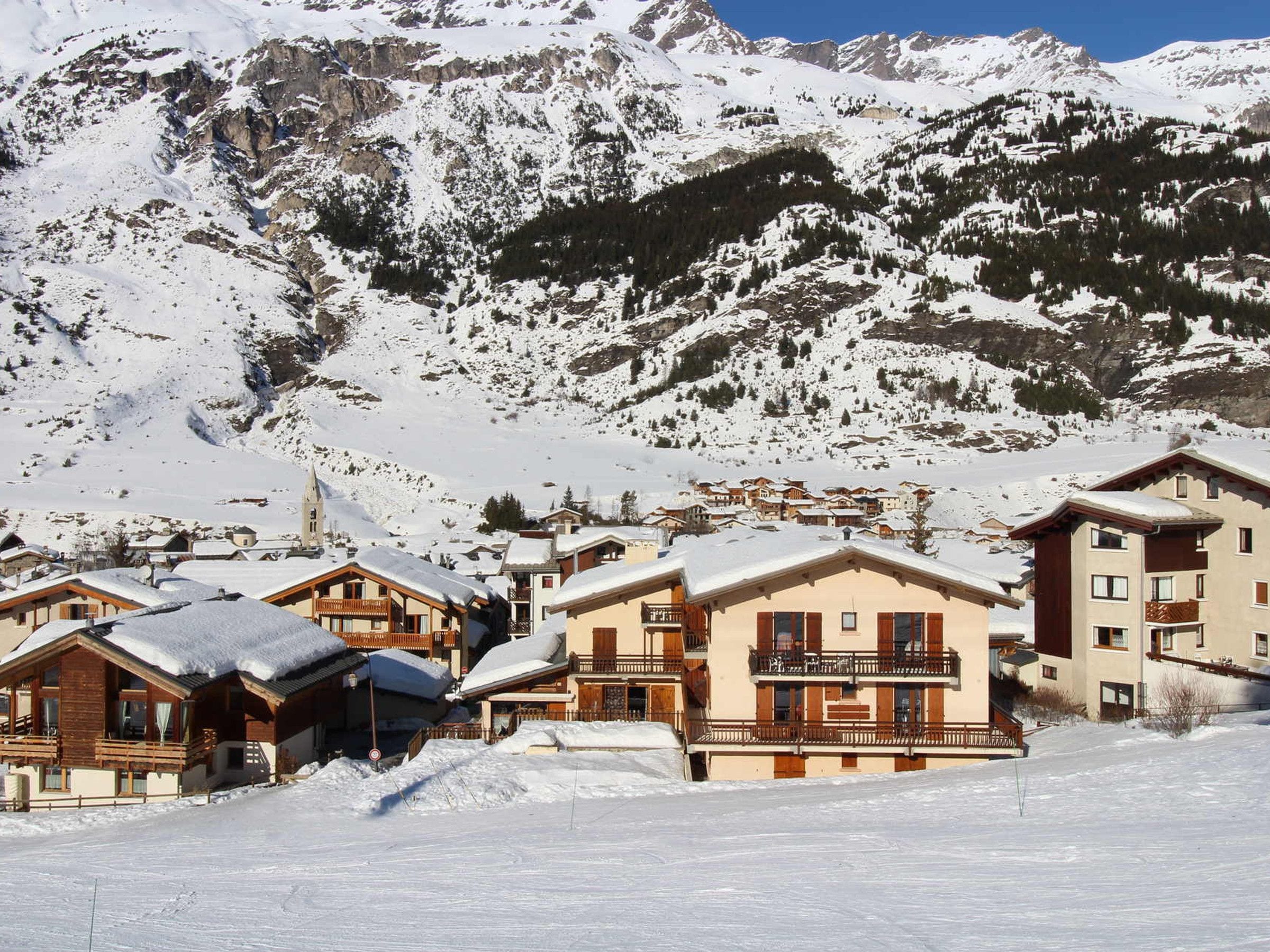 Un pueblo en la montaña cubierto de nieve con edificios tipo chalé, un telesilla y un cielo azul despejado.