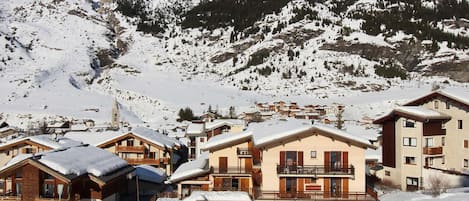 Un pueblo en la montaña cubierto de nieve con edificios tipo chalé, un telesilla y un cielo azul despejado.