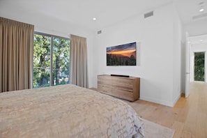 A bedroom with a large bed, a wooden dresser, a painting on the wall, and a sliding glass door leading to a balcony.