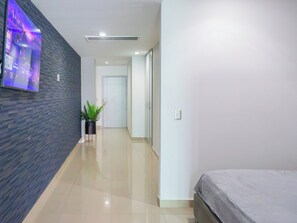Ein Hotelkorridor mit einem an der Wand befestigten Flachbildfernseher, einem Topf mit Pflanze und einem Bett mit grauer Bettdecke.