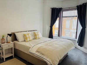 Quarto com cama, mesa de cabeceira, luminária e janela com cortinas.