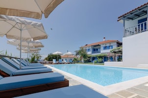 A pool area with lounge chairs and umbrellas, a clear sky, and a building with blue shutters.