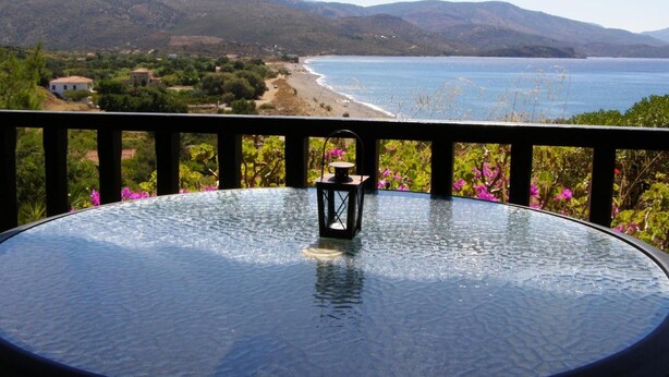 A glass table with a lantern on it, overlooking a beach and mountains. - Marvina (Chios)