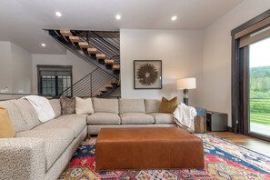 Ein modernes Wohnzimmer mit einem Ecksofa, einem braunen Hocker, einem gemusterten Teppich, einer Treppe und einem großen Fenster.